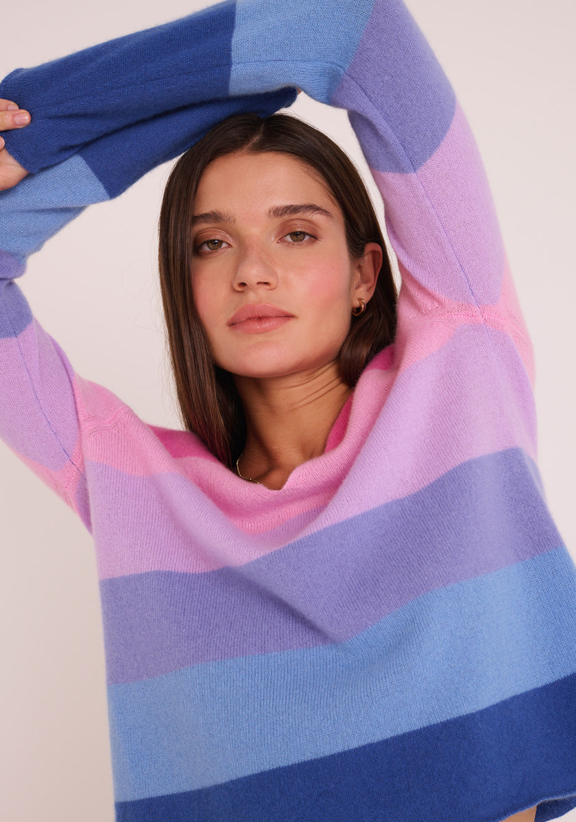 Eine junge Frau mit glattem braunem Haar trägt einen gemütlichen, gestreiften Feinstrickpullover in Blau, Lila und Rosa von Princess Goes Hollywood. Sie posiert mit einem erhobenen Arm vor einem schlichten, hellen Hintergrund und blickt ruhig in die Kamera.