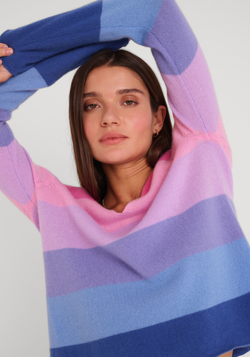 Eine junge Frau mit glattem braunem Haar posiert mit erhobenem Arm und trägt einen gemütlichen Princess Goes Hollywood Kaschmir Pullover mit kräftigen blauen, lila und rosa Streifen. Sie blickt neutral in die Kamera vor einem hellgrauen Hintergrund.