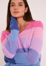 Eine Frau mit langen braunen Haaren trägt einen Princess Goes Hollywood Feinstrickpullover mit horizontalen rosa, lavendelfarbenen, blauen und lila Streifen. Sie stützt ihre Hand mit dem blauen Ärmel auf ihre Wange und blickt ruhig in die Kamera vor einem hellen Hintergrund.