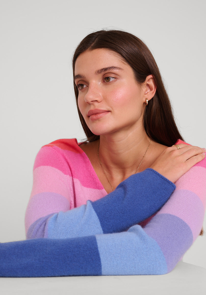 Eine Frau mit glattem braunem Haar sitzt an einem Tisch und trägt einen weichen Princess Goes Hollywood Feinstrickpullover mit V-Ausschnitt in Rosa, Lila und Blau. Sie stützt ihren Arm auf den Tisch und blickt ruhig zur Seite.