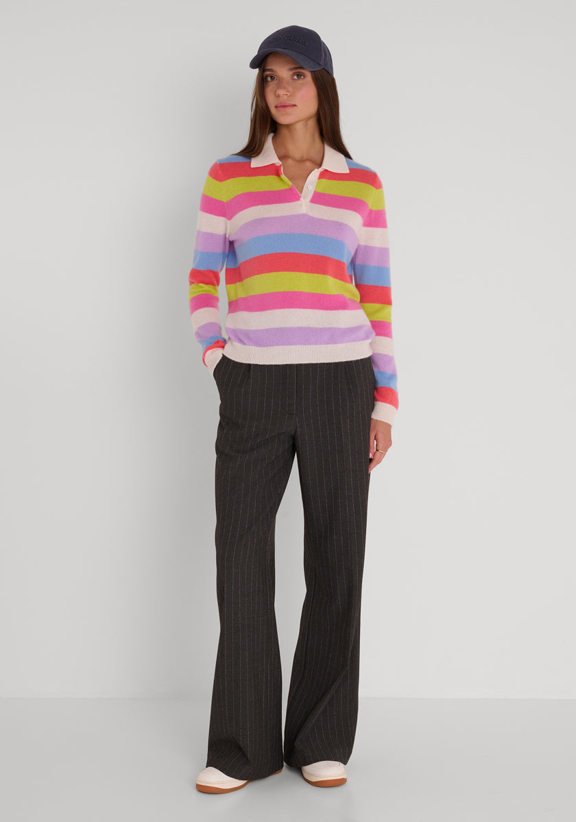 Eine Frau steht an einer schlichten Wand. Sie trägt einen bunt gestreiften Feinstrickpullover von Princess Goes Hollywood, eine marineblaue Baseballkappe, eine dunkelgraue Hose mit weitem Bein in Nadelstreifen und weiße Turnschuhe. Sie blickt nach vorne und hat die Hände in den Taschen.