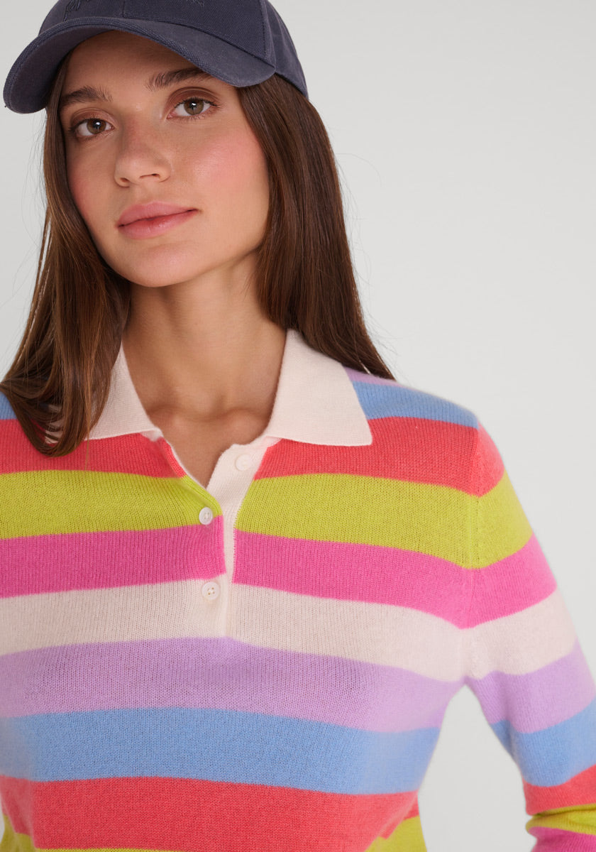 Eine Frau mit langen braunen Haaren trägt eine marineblaue Mütze und einen Princess Goes Hollywood Feinstrickpullover mit Polokragen, der horizontale Streifen in Rot, Rosa, Lila, Blau, Weiß und Lindgrün aufweist. Sie blickt leicht nach links vor einem hellen Hintergrund.