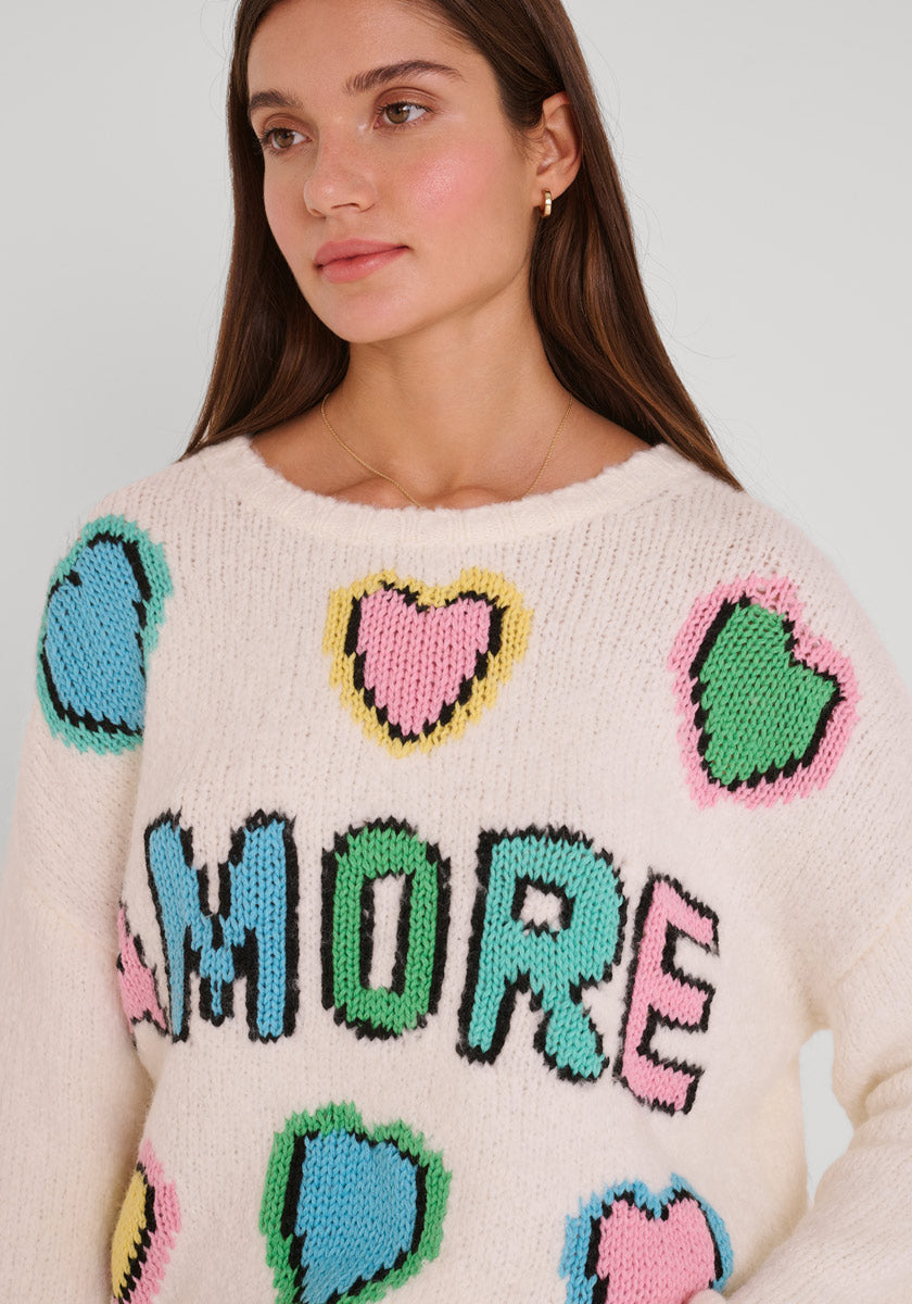 Eine Frau mit langen braunen Haaren trägt einen Princess Goes Hollywood Wollmix Grobstrickpullover mit bunten Amore-Intarsien und einem handgestrickten Amore-Logo. Sie posiert ruhig, mit kleinen Ohrringen und einer zarten Halskette vor einem schlichten Hintergrund.