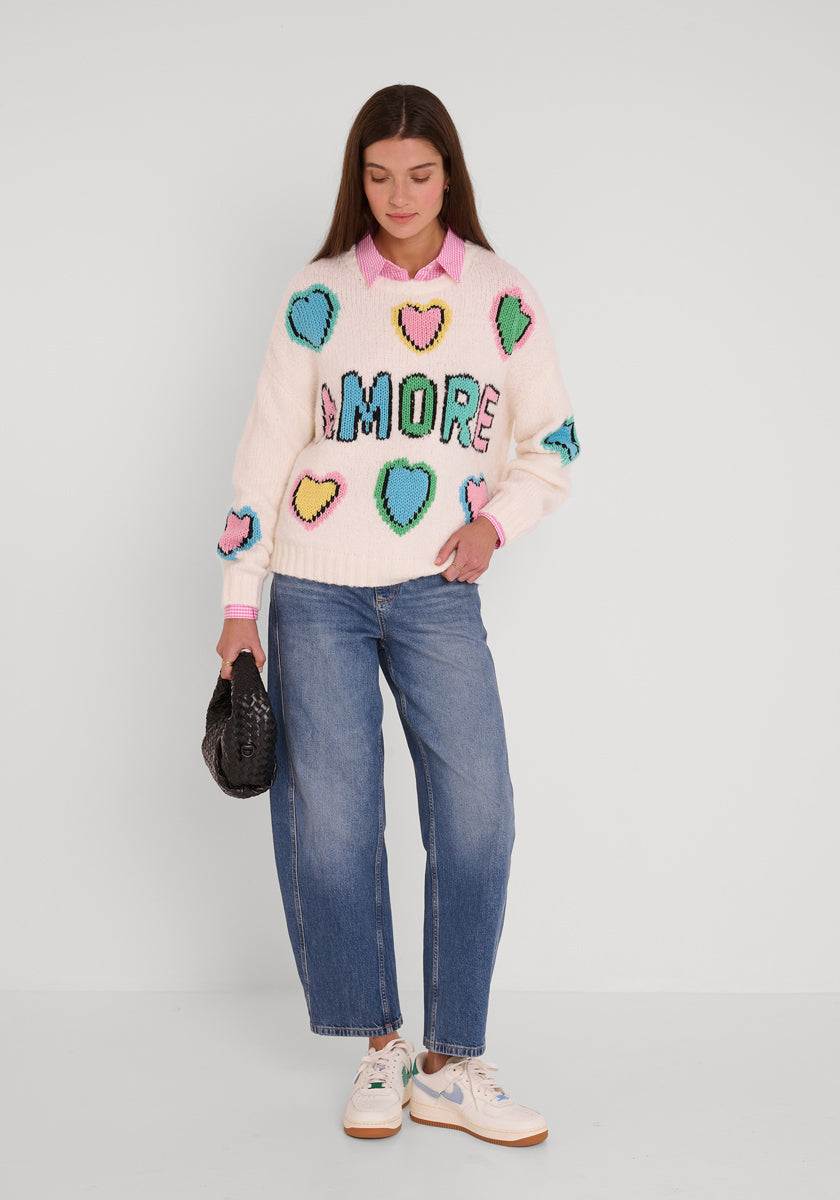 Eine Frau blickt nach vorne vor einem schlichten weißen Hintergrund und lächelt sanft. Sie trägt einen Princess Goes Hollywood Wollmix Grobstrickpullover mit bunten Herzen und handgestricktem Amore Schriftzug, blaue Jeans, weiße Turnschuhe und hält eine kleine schwarze Handtasche.