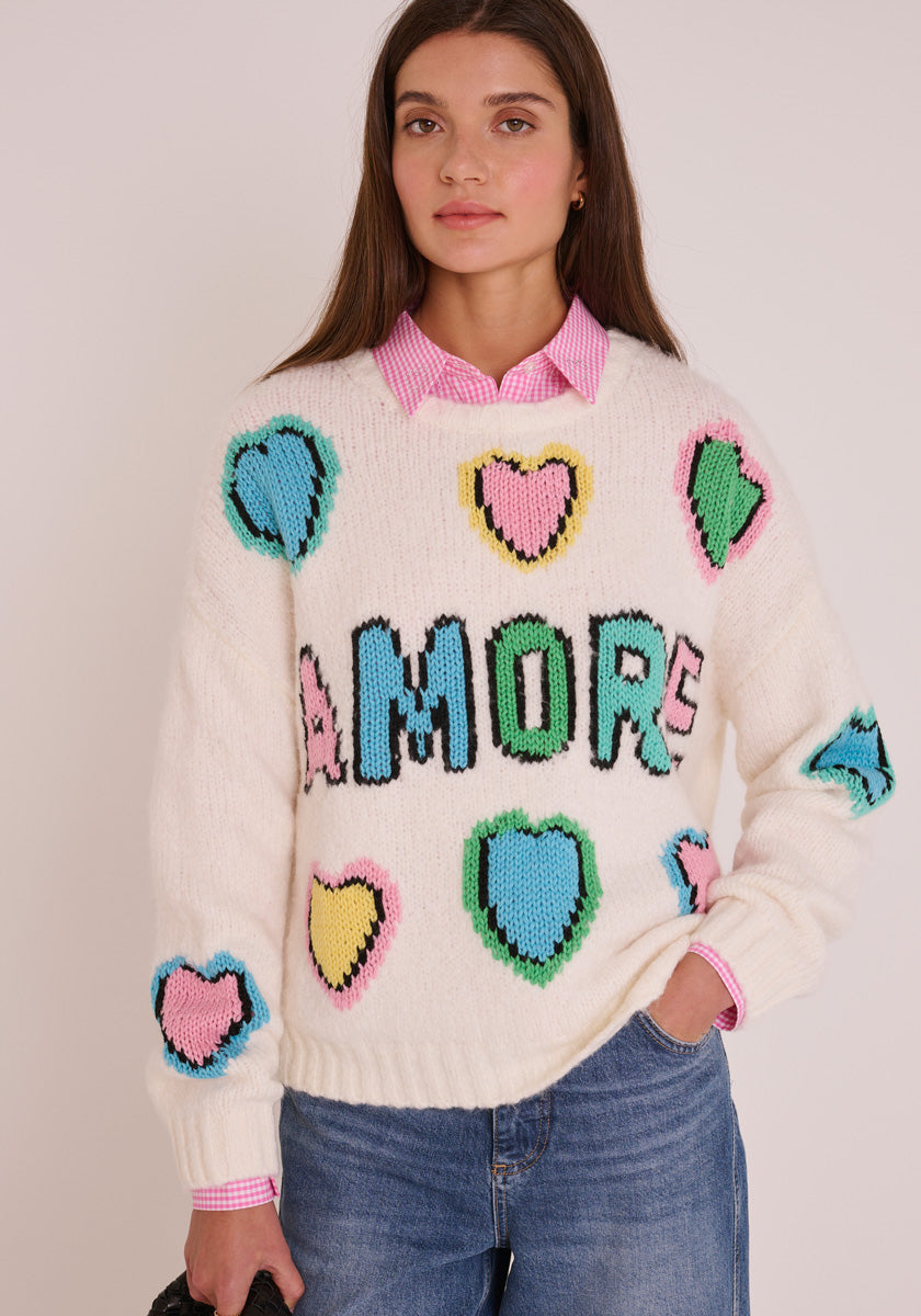 Eine Frau mit glattem braunem Haar trägt einen Princess Goes Hollywood Wollmix Grobstrickpullover mit Herz Intarsien und handgestricktem Amore Schriftzug, der über einem pinkfarbenen, karierten Hemd und blauen Jeans liegt, und posiert mit einer Hand in ihrer Tasche.