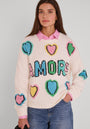 Eine Frau mit langen braunen Haaren trägt einen weißen Pullover aus einem Wollgemisch mit großen bunten gestrickten Herzen und dem Schriftzug "AMORE". Darunter trägt sie eine blaue Jeans und ein rosa kariertes Hemd.