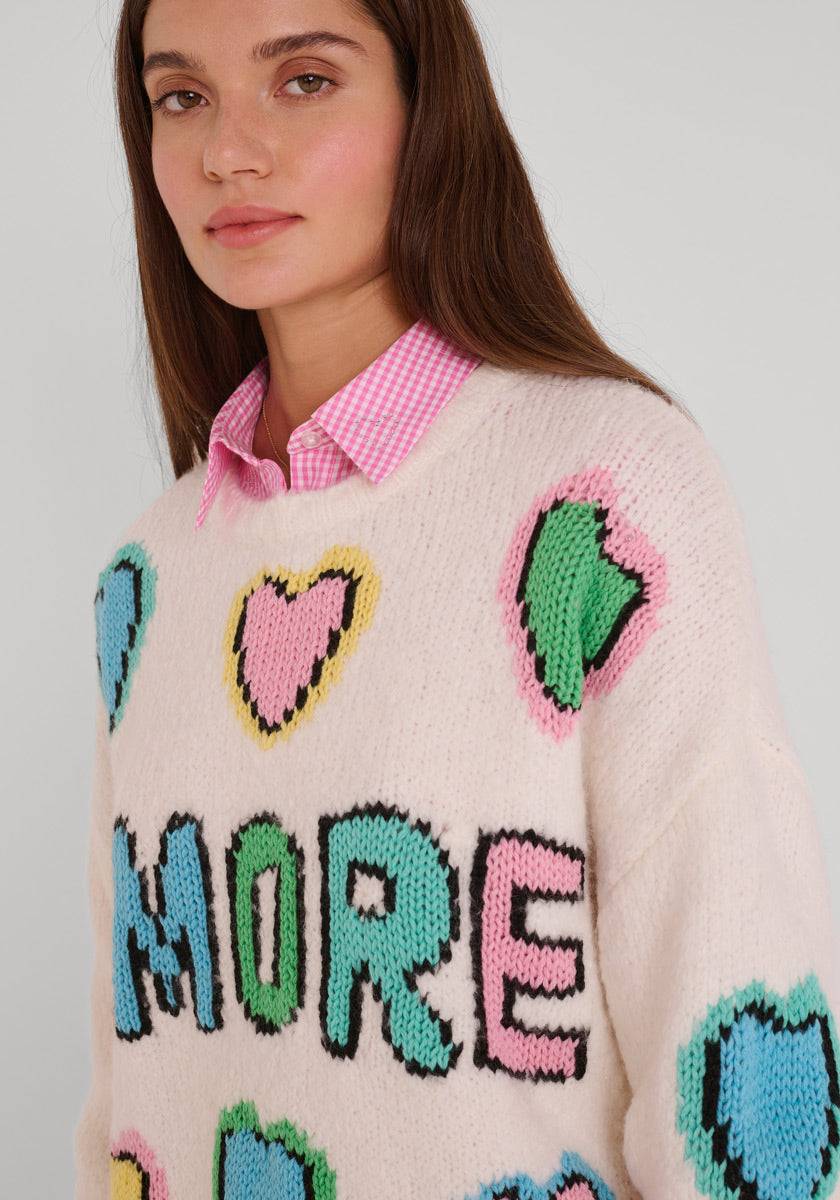 Eine Frau mit langen braunen Haaren trägt unter einem Wollmix-Grobstrickpullover mit großen bunten Herzen und einem auffälligen, mehrfarbigen, handgestrickten Amore-Schriftzug ein pinkfarbenes, kariertes Princess Goes Hollywood-Kragenhemd, während sie zur Seite schaut.