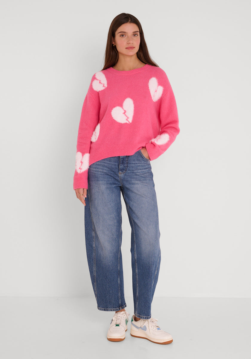 Eine Frau mit langen braunen Haaren und einem neutralen Gesichtsausdruck steht vor einer weißen Wand. Sie trägt einen Wollpullover mit gebrochenen Herzen von Princess Goes Hollywood, lässige Bluejeans und weiße Turnschuhe mit pastellfarbenen Akzenten. Ihre linke Hand steckt in ihrer Tasche.