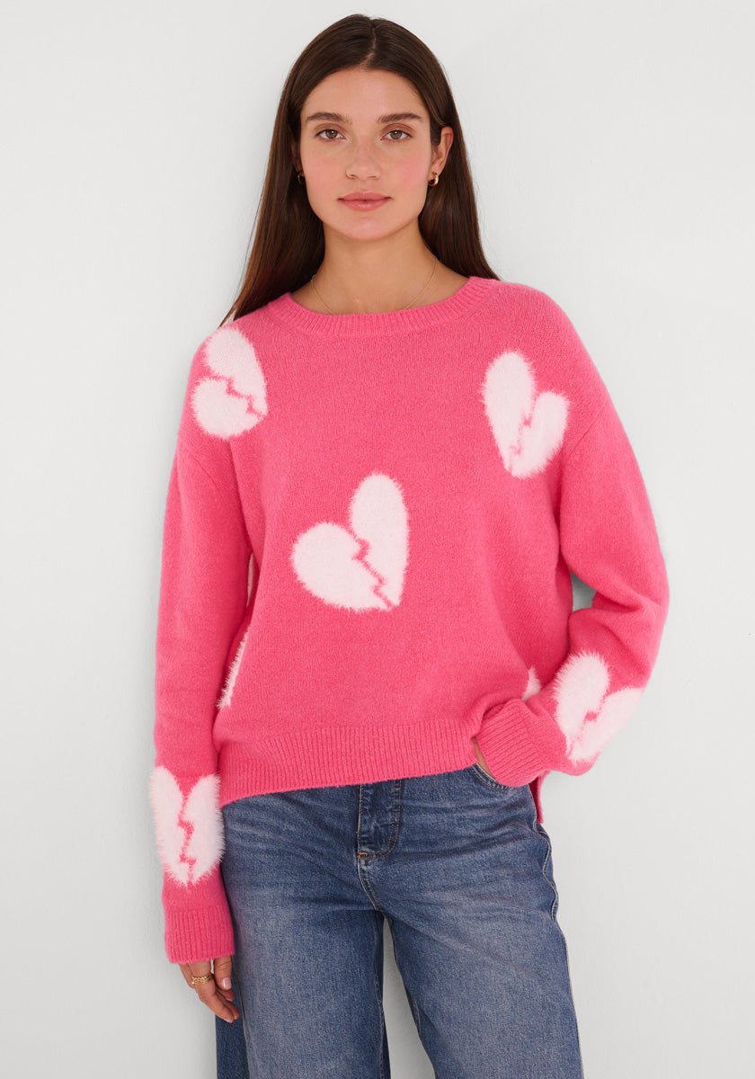Eine Frau mit langen braunen Haaren trägt einen rosa Princess Goes Hollywood Pullover aus Wollmix mit Rundhalsausschnitt und großen weißen Broken-Heart-Designs. Sie steht vor weißem Hintergrund, eine Hand in der Jeans, mit neutraler Miene.