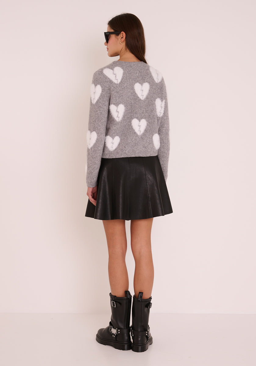 Eine Frau steht vor einer hellen Wand und trägt eine graue Strickjacke aus einer Wollmischung mit großen Herzmotiven, einen schwarzen Faltenminirock aus Kunstleder, schwarze Bikerstiefel mit Schnallen und eine Sonnenbrille.