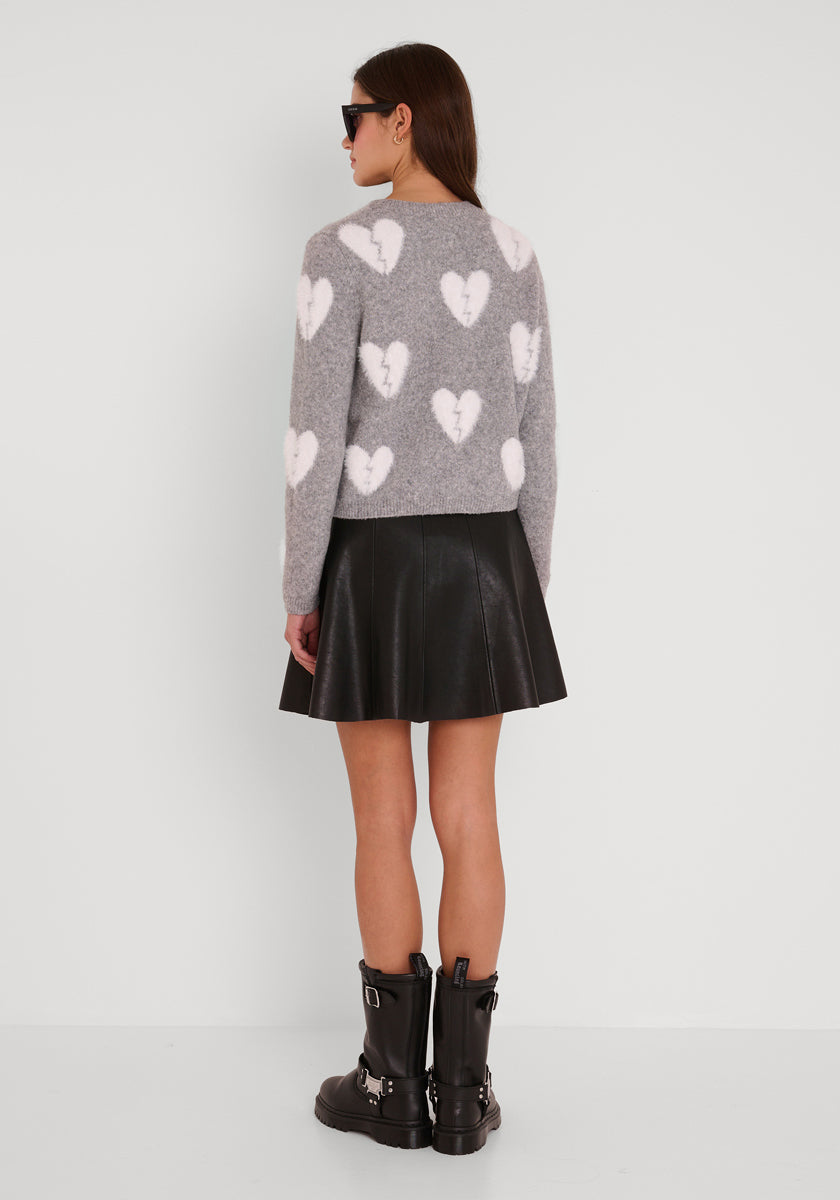 Eine Frau trägt eine graue Strickjacke mit weißen Herz-Motiven von Princess Goes Hollywood, einen schwarzen plissierten Lederrock, schwarze Stiefeletten mit Schnallen und Sonnenbrille. Ihr braunes Haar fällt locker vor weißen Hintergrund.