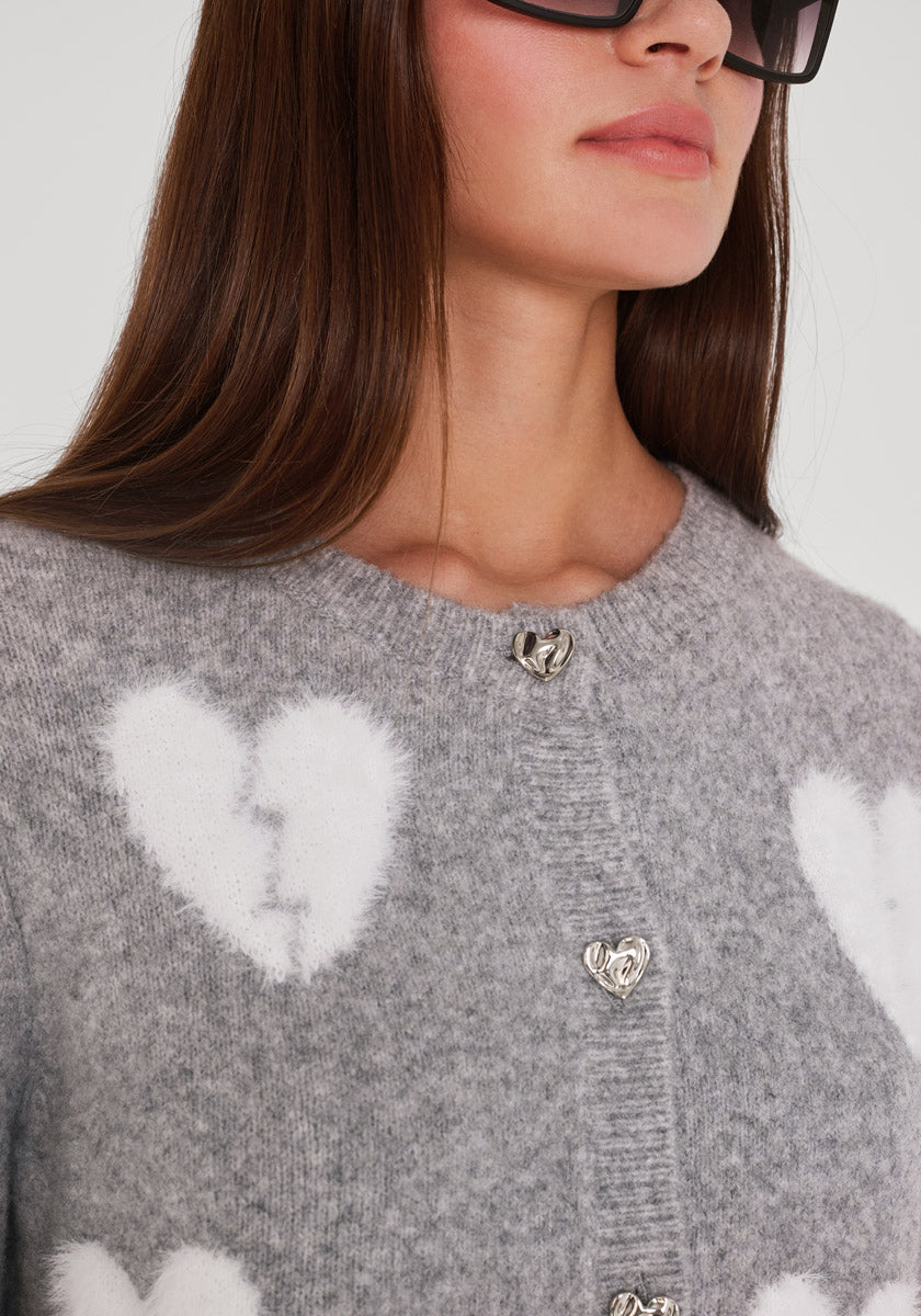 Eine Frau mit langen braunen Haaren trägt eine übergroße schwarze Sonnenbrille und eine graue Princess Goes Hollywood Wollmix Strickjacke mit weißen Herzmotiven und silbernen herzförmigen Knöpfen, die in einem Bildausschnitt gezeigt wird, der die Struktur des Pullovers hervorhebt.