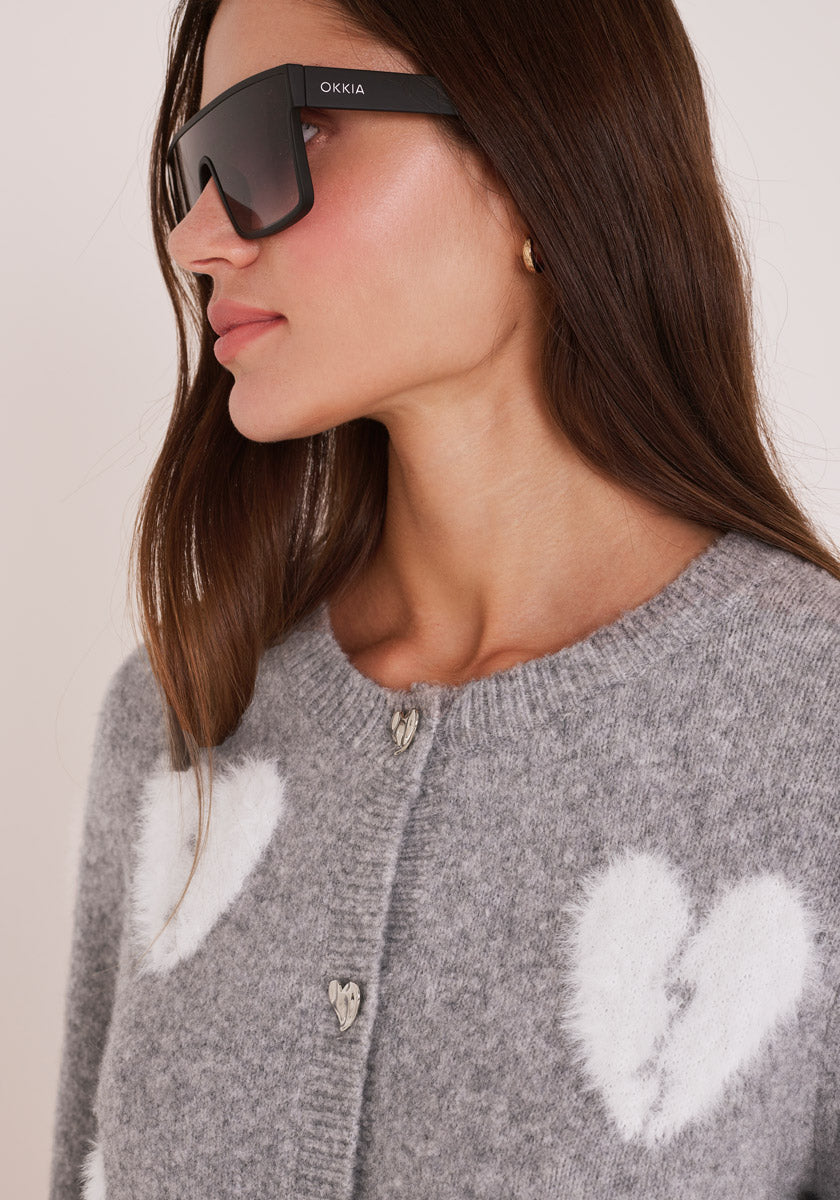 Eine Frau mit langen braunen Haaren trägt eine große schwarze Sonnenbrille und eine Princess Goes Hollywood Wollmix Strickjacke mit weißen Herzmotiven, herzförmigen Knöpfen und kleinen goldenen Ohrringen; ihr Kopf ist leicht nach links gedreht.