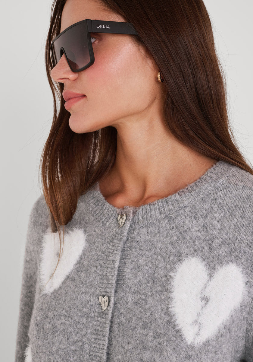 Eine Frau mit langen braunen Haaren trägt eine übergroße schwarze Sonnenbrille und eine graue Princess Goes Hollywood Wollmix Strickjacke mit großen weißen Herzmotiven und herzförmigen Knöpfen. Sie blickt nach links und trägt natürliches Make-up und einen goldenen Ohrring.