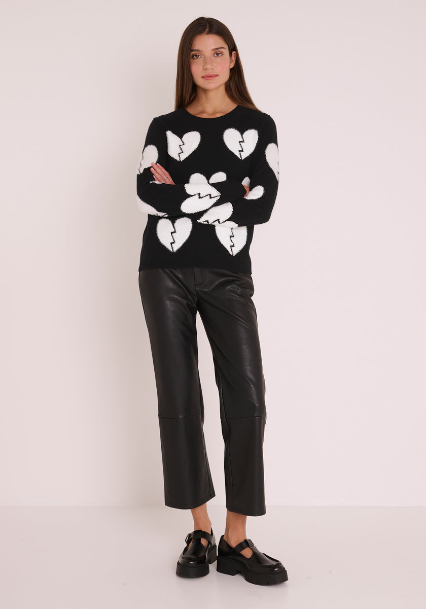 Eine Frau steht vor einer weißen Wand in einem Princess Goes Hollywood Wollmix-Pullover mit Rundhalsausschnitt und großem weißem Herz-Motiv, gepaart mit einer schwarzen Kunstlederhose und klobigen Schuhen. Ihr Haar ist lang und glatt; sie hat die Arme verschränkt und einen neutralen Blick.