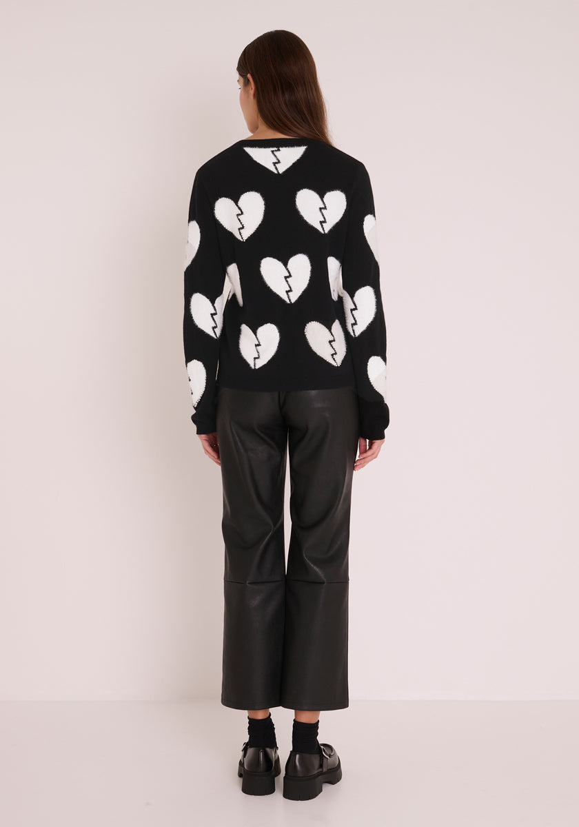 Eine Person mit langen braunen Haaren steht vor einer weißen Wand. Sie trägt einen gewagten Princess Goes Hollywood Wollmix-Pullover mit Rundhalsausschnitt und großem weißen Herz-Motiv, gepaart mit einer schwarzen, weit geschnittenen Lederhose und schwarzen Schuhen für einen modernen Look.