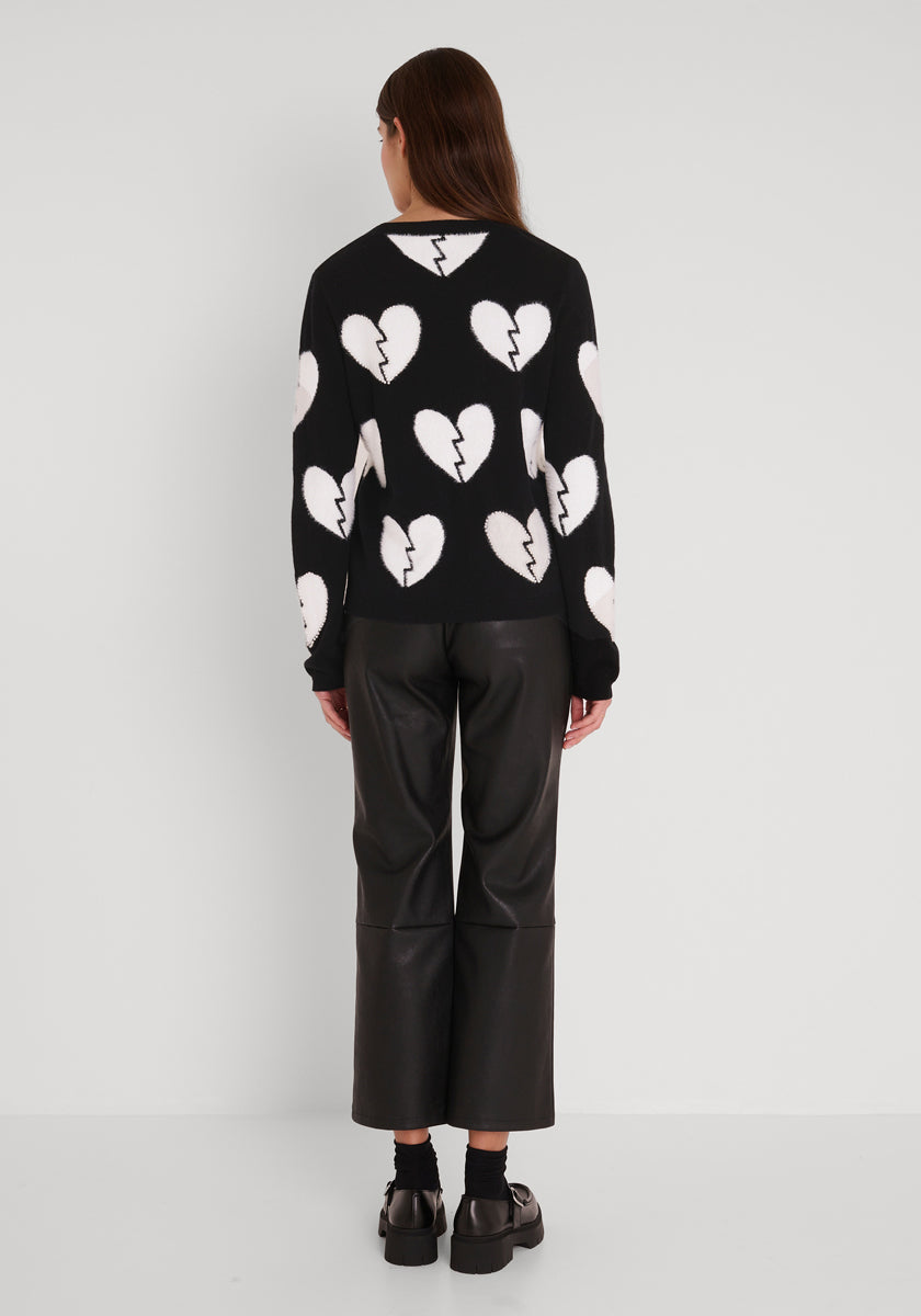 Eine Person mit langen braunen Haaren, von hinten gesehen, trägt einen Wollpullover mit großen gebrochenen Herzen, eine schwarze, weit geschnittene Hose und schwarze Schuhe mit Socken vor einem weißen Hintergrund.