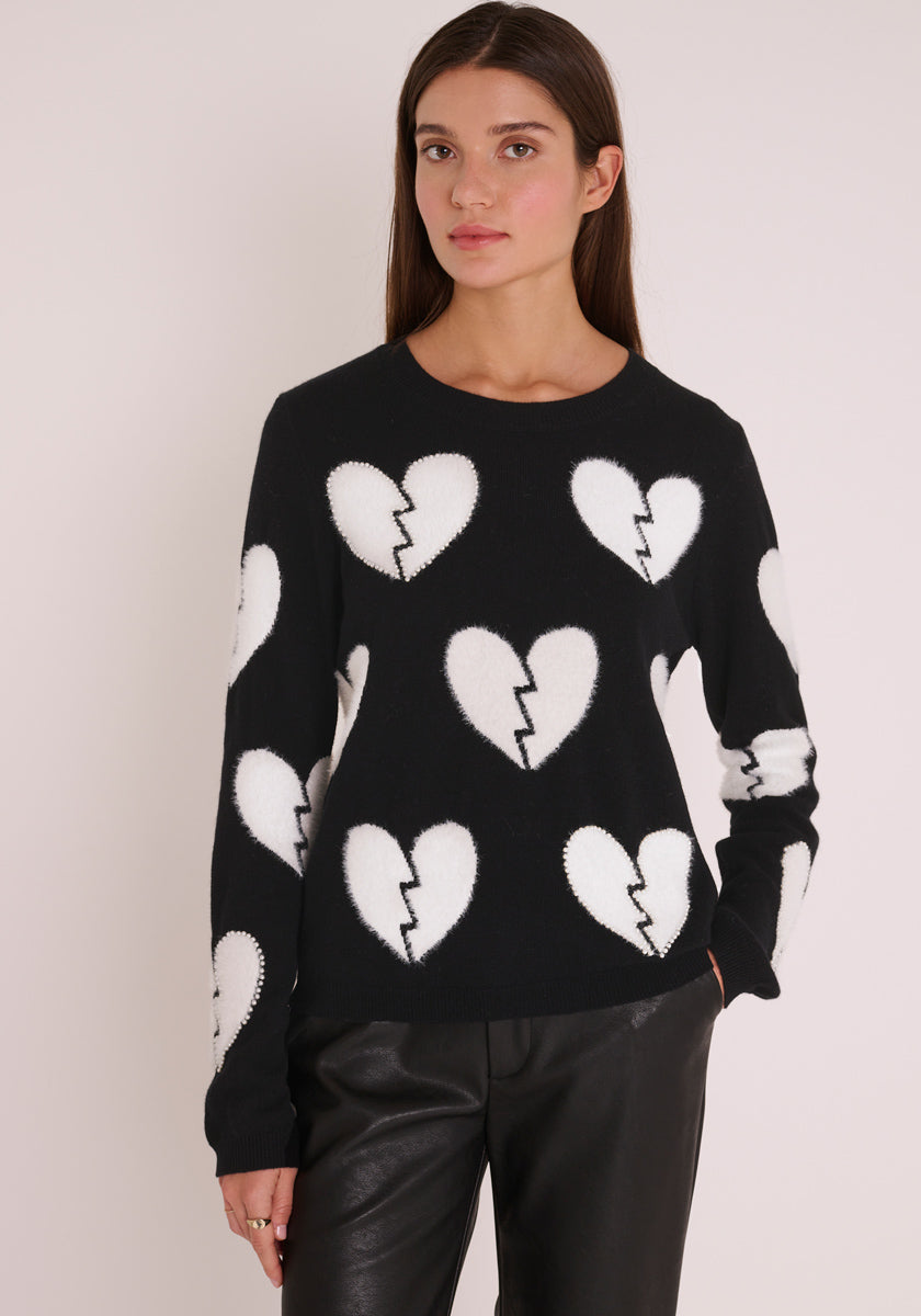 Eine Frau mit langen braunen Haaren trägt einen Wollmix-Pullover mit Rundhalsausschnitt von Princess Goes Hollywood, geschmückt mit großen gebrochenen Herz-Motiven, sowie schwarzen Hosen. Sie steht vor hellem Hintergrund und blickt neutral nach vorne.