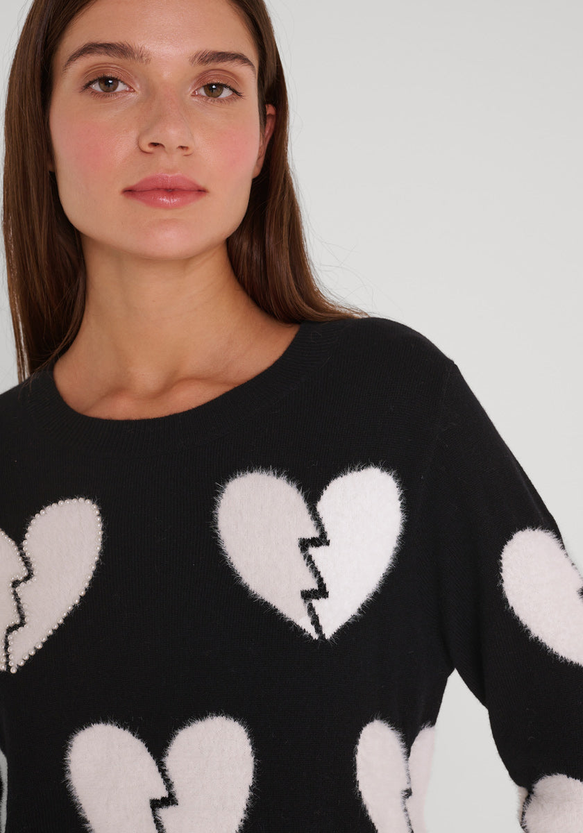 Eine Frau mit langen braunen Haaren und natürlichem Make-up trägt einen Pullover aus einer Wollmischung von Princess Goes Hollywood mit großen, weißen, gebrochenen Herzmotiven und Rundhalsausschnitt. Sie blickt neutral in die Kamera vor einem schlichten hellgrauen Hintergrund.