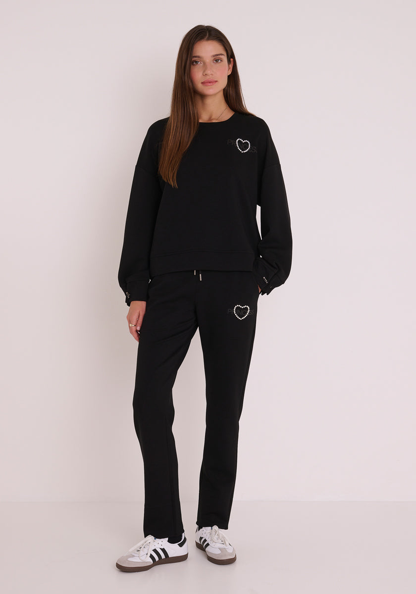 Eine Frau mit langen braunen Haaren trägt ein schwarzes Princess Goes Hollywood-Sweatshirt und dazu passende Jogginghosen, beide mit einem kleinen weißen Herz. Sie steht in weißen Adidas-Turnschuhen mit schwarzen Streifen vor einem schlichten weißen Hintergrund.