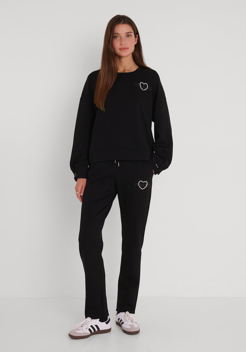 Eine Frau mit langen braunen Haaren steht vor einer weißen Wand. Sie trägt ein lockeres Princess Goes Hollywood Sweatshirt Damen mit Rundhalsausschnitt und eine dazu passende Jogginghose, auf der jeweils ein kleines weißes Herz abgebildet ist. Dazu trägt sie weiße Turnschuhe mit schwarzen Streifen.