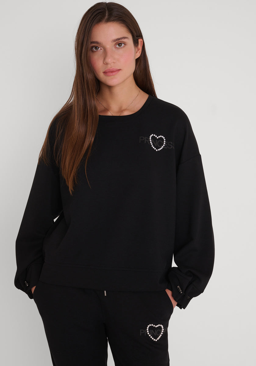 Eine junge Frau mit langen braunen Haaren trägt ein locker sitzendes schwarzes Princess Goes Hollywood-Sweatshirt und eine passende Hose, beide mit einem kleinen weißen Perlenherz auf der linken Seite. Sie steht vor einem schlichten weißen Hintergrund und schaut neutral in die Kamera.