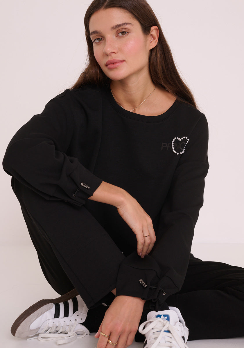 Eine Frau mit langen braunen Haaren sitzt auf dem Boden vor einem weißen Hintergrund. Sie trägt ein schwarzes Princess Goes Hollywood-Sweatshirt mit einer Herzverzierung an der Schulter, eine passende schwarze Hose und weiße Adidas-Turnschuhe und blickt neutral in die Kamera.