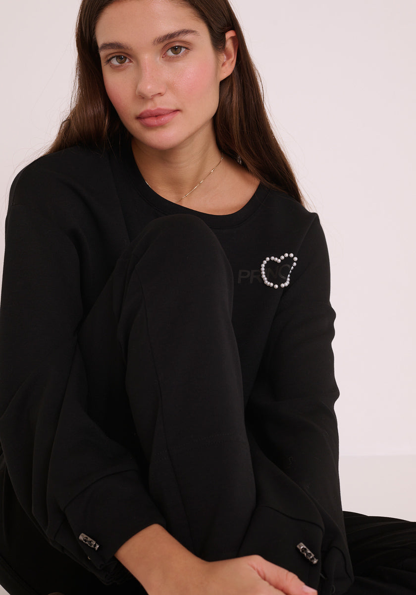 Eine Frau mit langen braunen Haaren und heller Haut sitzt mit einem Knie nach oben und trägt ein schwarzes Princess Goes Hollywood-Sweatshirt mit einem PRINCESS-Logo und einer herzförmigen Perlenbrosche auf der Brust. Sie lächelt sanft vor einem cremefarbenen Hintergrund.