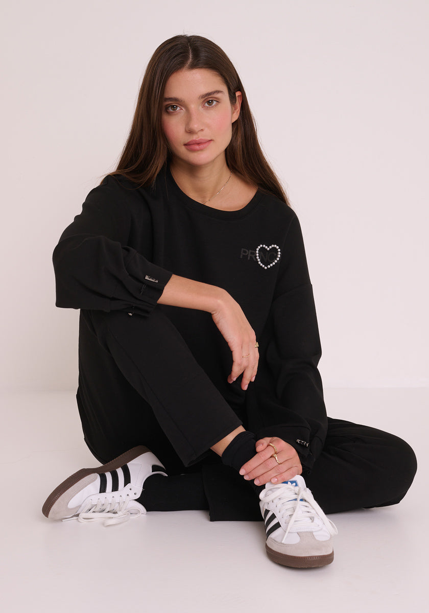 Eine Frau mit langen braunen Haaren sitzt auf dem Boden vor einem weißen Hintergrund. Sie trägt ein schwarzes, langärmeliges Princess Goes Hollywood-Top mit Herz und PRINCESS-Logo, eine Sweathose mit elastischem Bund und weiße Adidas-Turnschuhe mit schwarzen Streifen.
