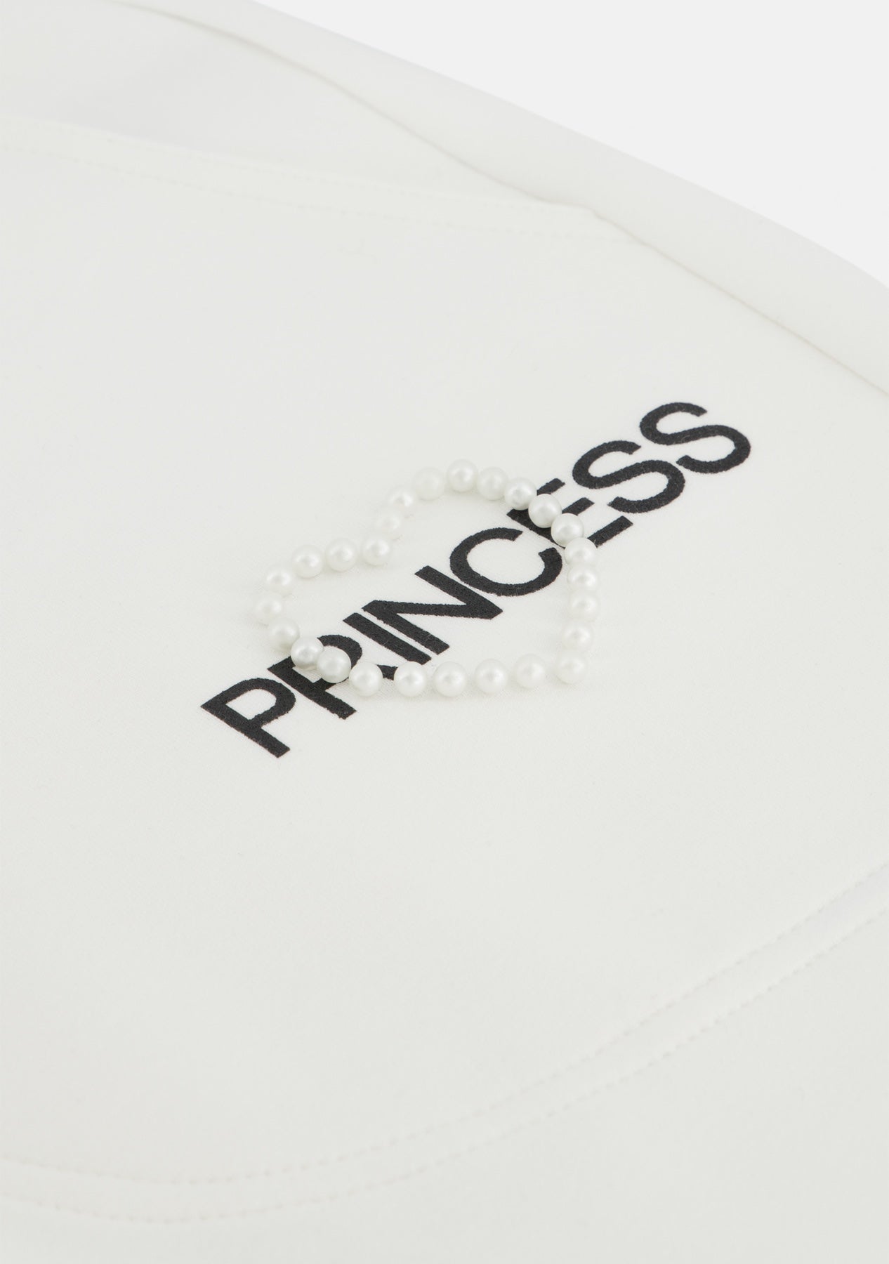 Eine Nahaufnahme des weißen Stoffes von Princess Goes Hollywood zeigt das aufgestickte schwarze PRINCESS-Logo, das von einem Perlenarmband in Herzform gekrönt wird. Der weiche, minimale Hintergrund erinnert an eine Sweathose mit elastischem Bund.