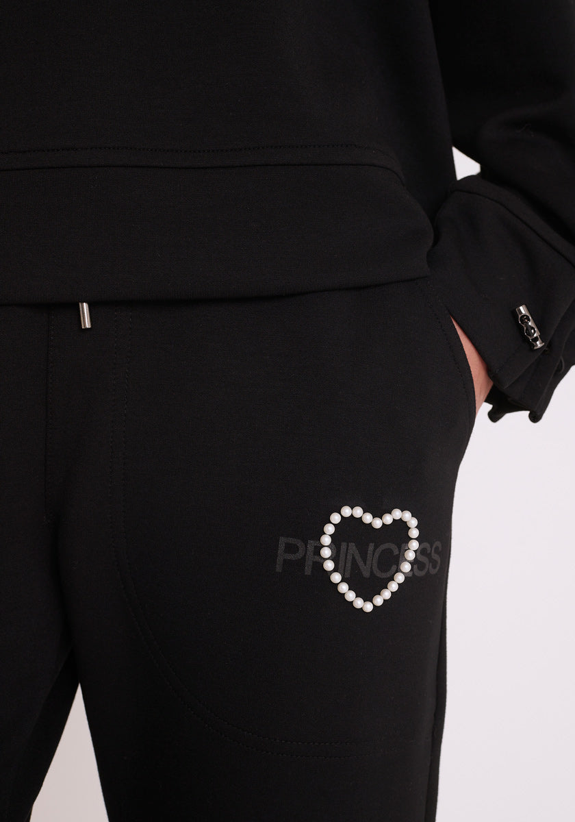 Nahaufnahme einer Person, die eine schwarze Princess Goes Hollywood Sweathose mit einem elastischen Bund und einem PRINCESS-Logo mit Perlenherz trägt. Ihre Hand ist in der Nähe der Tasche, und ein Teil eines schwarzen Sweatshirts mit einem silbernen Reißverschluss ist sichtbar.