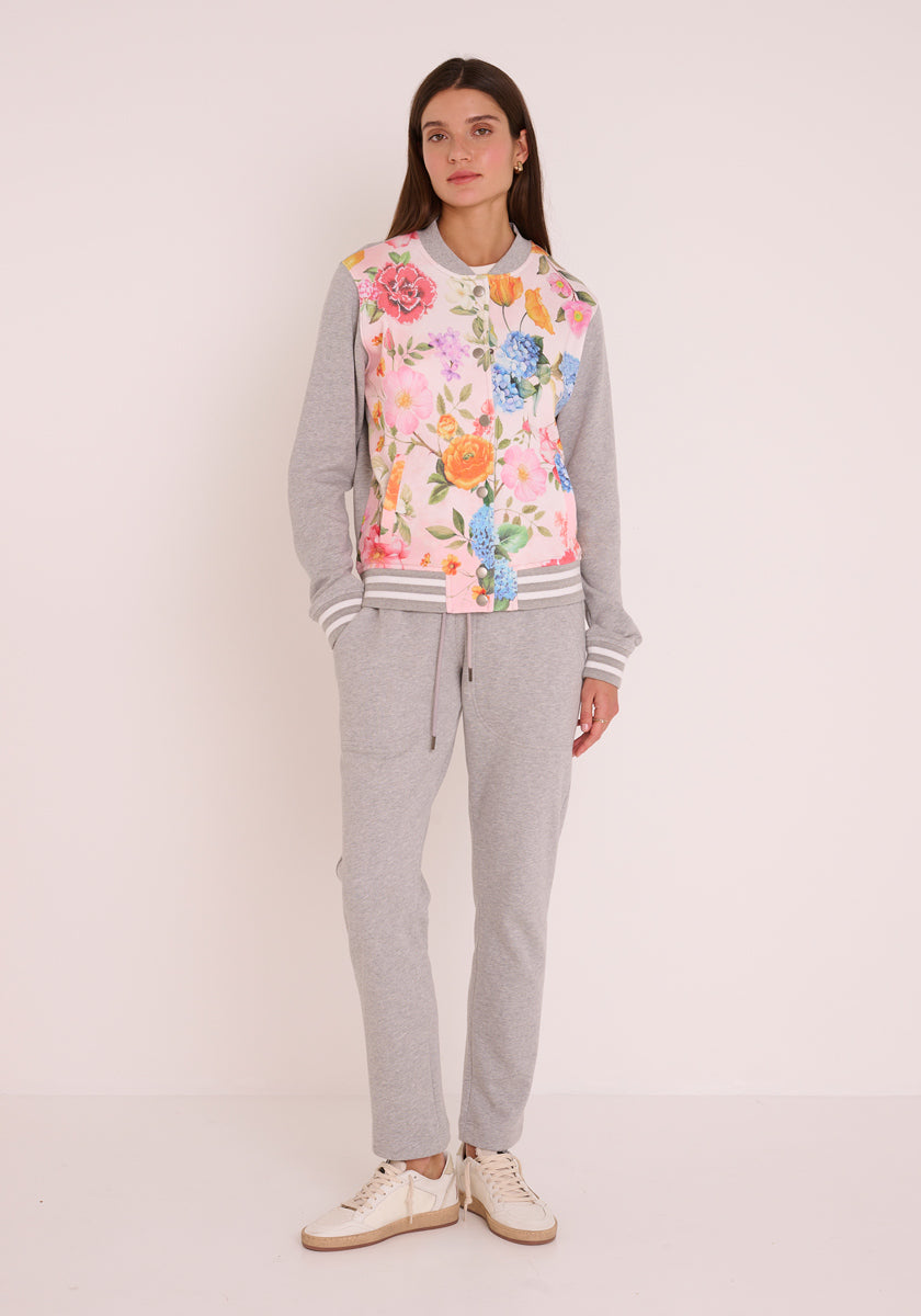 Eine Frau mit langen braunen Haaren und einem neutralen Gesichtsausdruck trägt eine graue Princess Goes Hollywood-Sweatjacke und Turnschuhe. Ihr Look im College-Stil besteht aus einem Sweatshirt mit grauen Ärmeln und einem bunten Blumendruck auf der Vorderseite, der sich von einem schlichten hellen Hintergrund abhebt.