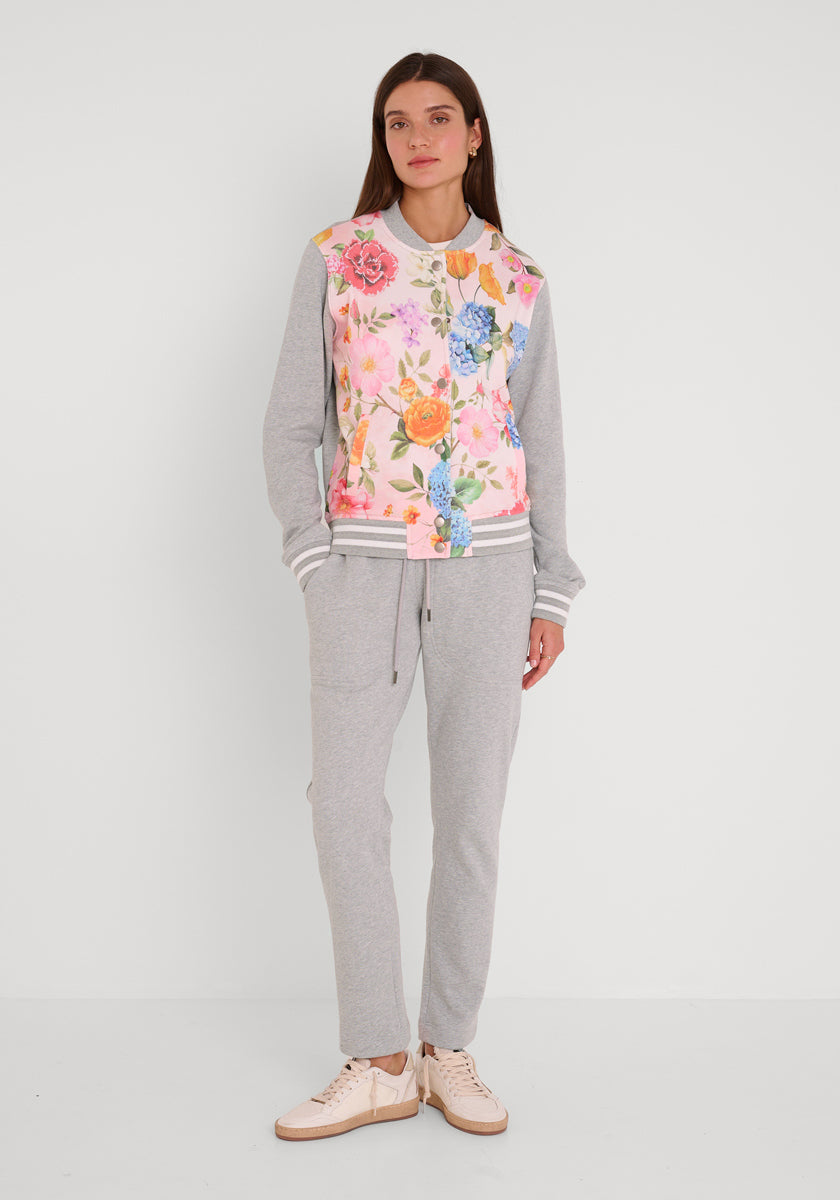 Eine Frau mit langen braunen Haaren und neutralem Gesichtsausdruck steht vor einem weißen Hintergrund. Sie trägt die hellgraue Baumwoll-Jogginghose Princess Goes Hollywood mit elastischem Bund, passende Turnschuhe und ein Sweatshirt mit Blumendruck.