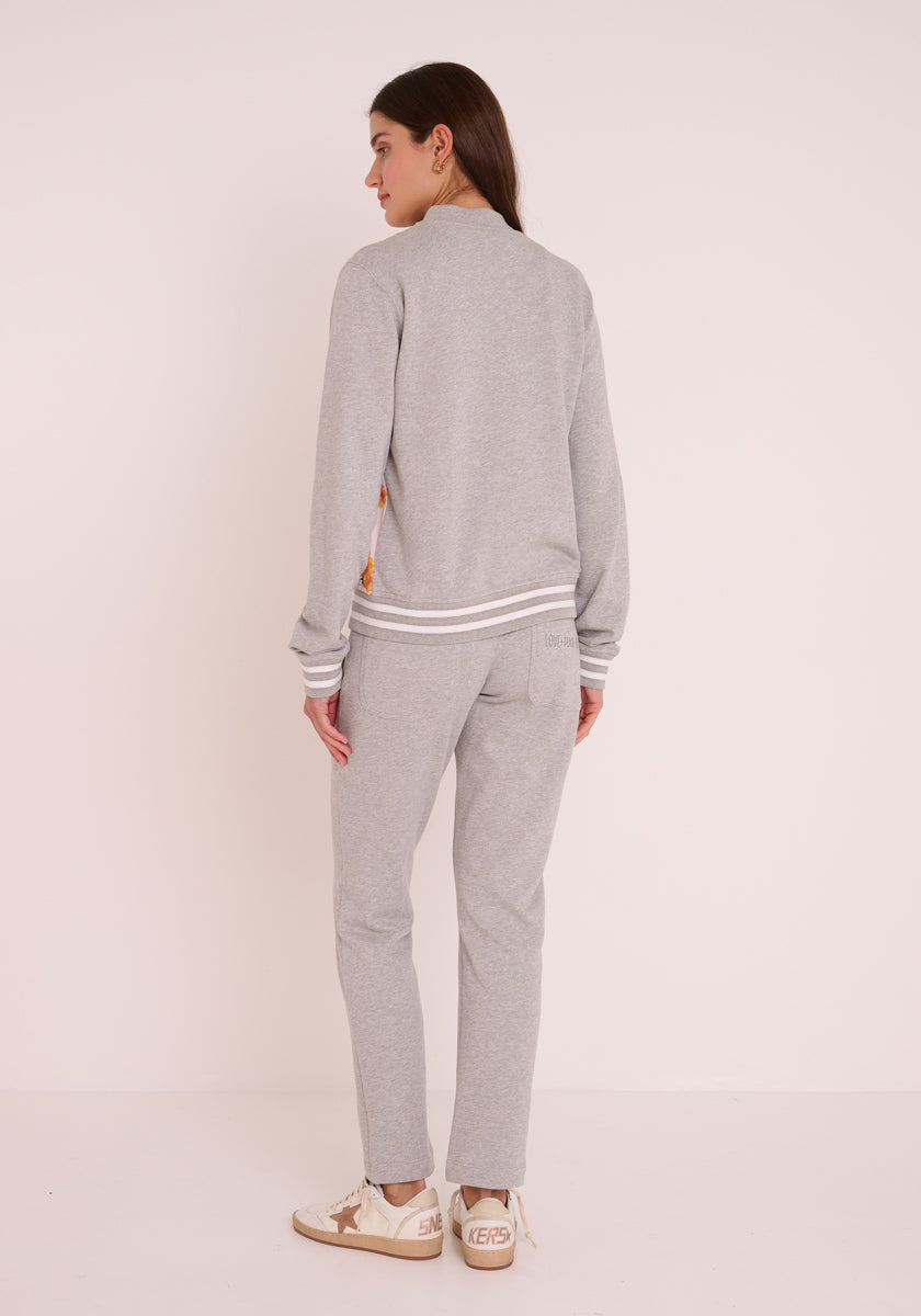 Eine Frau mit langen braunen Haaren trägt einen Princess Goes Hollywood Trainingsanzug aus hellgrauer Baumwolle mit elastischem Bund, weiß gestreiften Manschetten und Kragen sowie beigen Turnschuhen mit goldenen Sternchen. Sie steht mit dem Gesicht zur Seite vor einem schlichten, cremefarbenen Hintergrund.