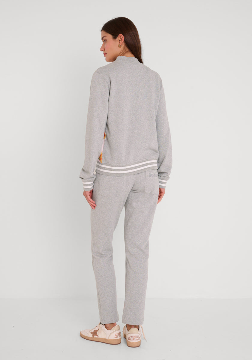 Eine Frau mit langen braunen Haaren trägt eine hellgraue Princess Goes Hollywood Sweatjacke im College-Stil mit weißem Besatz an den Ärmeln und am Bund, dazu beigefarbene Turnschuhe mit weißen Sohlen. Sie steht mit dem Gesicht zur Seite vor einem schlichten weißen Hintergrund.