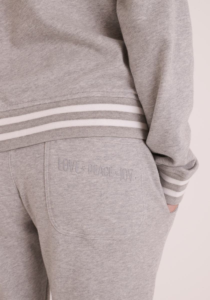 Eine Person trägt ein hellgraues Princess Goes Hollywood Baumwolle Sweatshirt und eine passende Sweathose mit elastischem Bund und weiß gestreiften Bündchen. Auf der Gesäßtasche ist &quot;LOVE - PEACE - JOY&quot; in grau aufgestickt. Die Hand befindet sich in der Tasche.