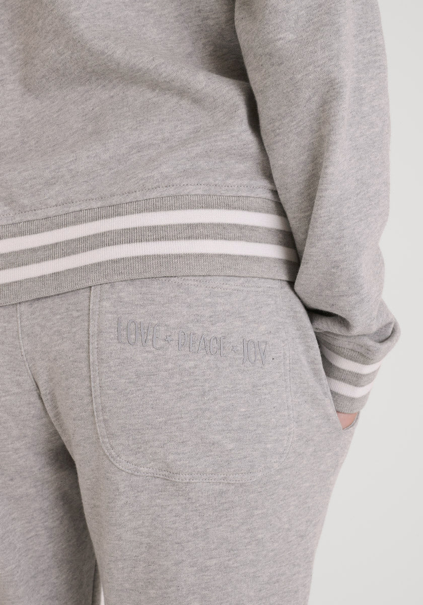 Nahaufnahme einer Person in einer hellgrauen Princess Goes Hollywood Sweatjacke und einer Jogginghose im College-Stil mit weiß und grau gestreiften Bündchen. "LOVE. FRIEDEN. JOY." ist auf die Gesäßtasche gestickt, während ihre Hand darin ruht.