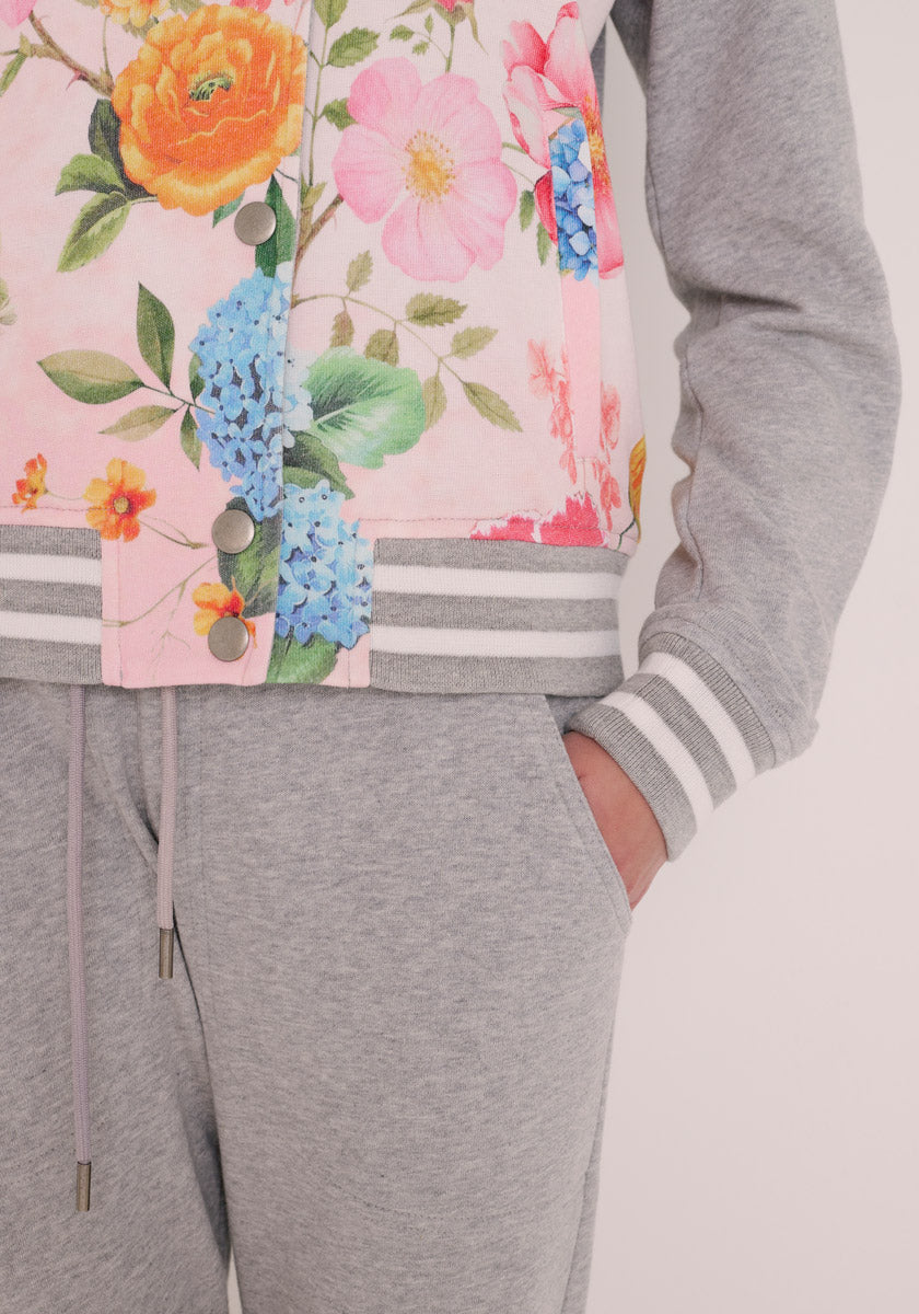 Eine Person in einer grauen Jogginghose und einer grauen Sweatjacke von Princess Goes Hollywood mit weiß gestreiften Bündchen und einem rosafarbenen Unterteil mit einem bunten Blumenaufdruck - orange, rosa und blaue Blumen - für einen entspannten College-Stil.