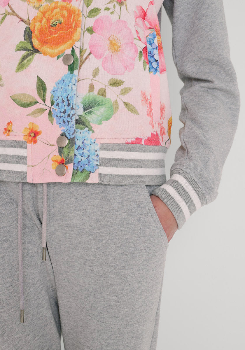 Eine Person trägt eine hellgraue Jogginghose und eine pastellrosa Princess Goes Hollywood College-Stil Sweatjacke mit einem bunten Blumenaufdruck (orange, rosa, blau), weiß und grau gestreiften Bündchen, Druckknöpfen und der rechten Hand in der Tasche.