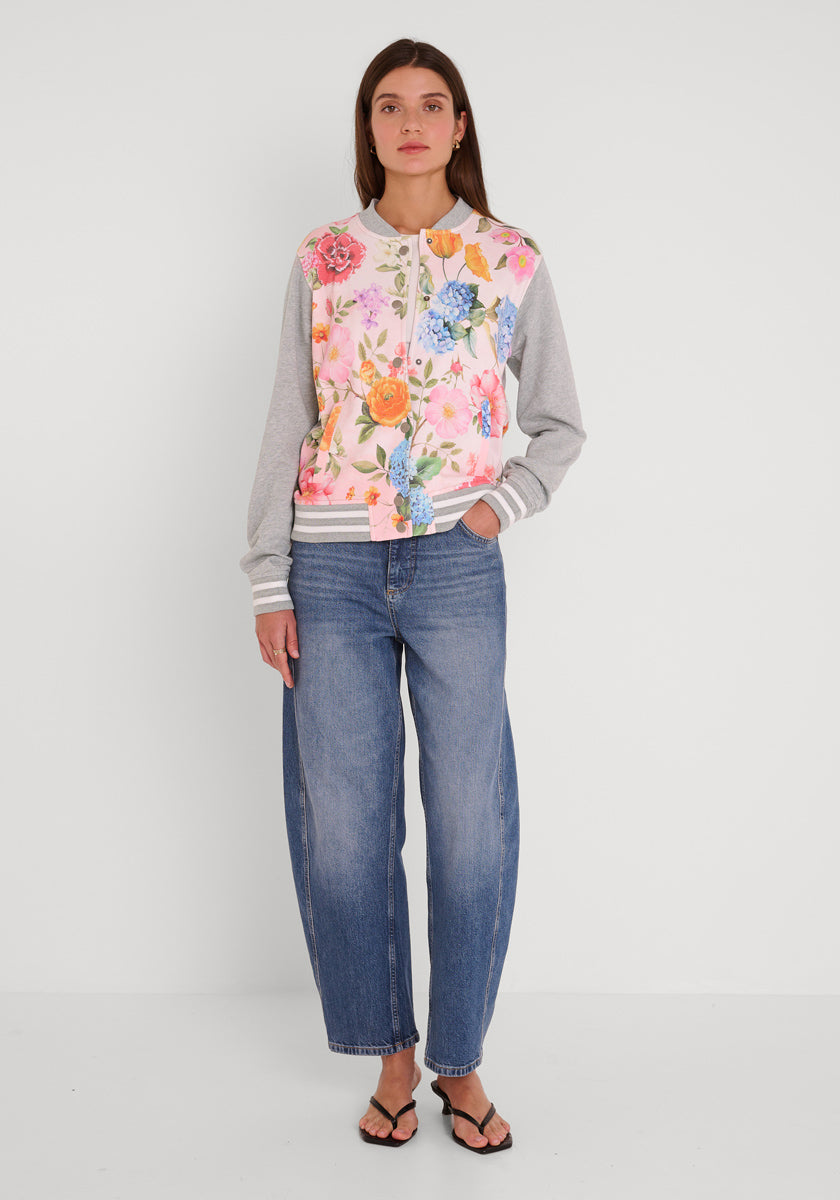 Eine Frau mit langen braunen Haaren steht vor einem weißen Hintergrund und trägt eine hellgraue und rosafarbene Blumen-Print-Sweatjacke von Princess Goes Hollywood mit bunten Blumenaufdrucken, weite Bluejeans und schwarze Flip-Flops und schaut geradeaus.