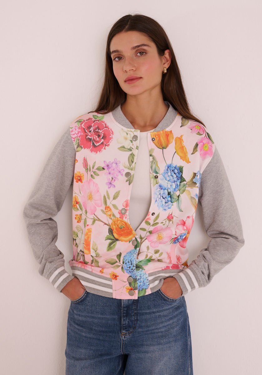 Eine Frau mit langen braunen Haaren trägt eine Princess Goes Hollywood Blumen-Print Bomberjacke im College-Stil mit grauen Ärmeln und gestreiften Bündchen, gepaart mit einer blauen Jeans. Sie steht an einer weißen Wand, die Hände in den Taschen, und schaut ruhig in die Kamera.