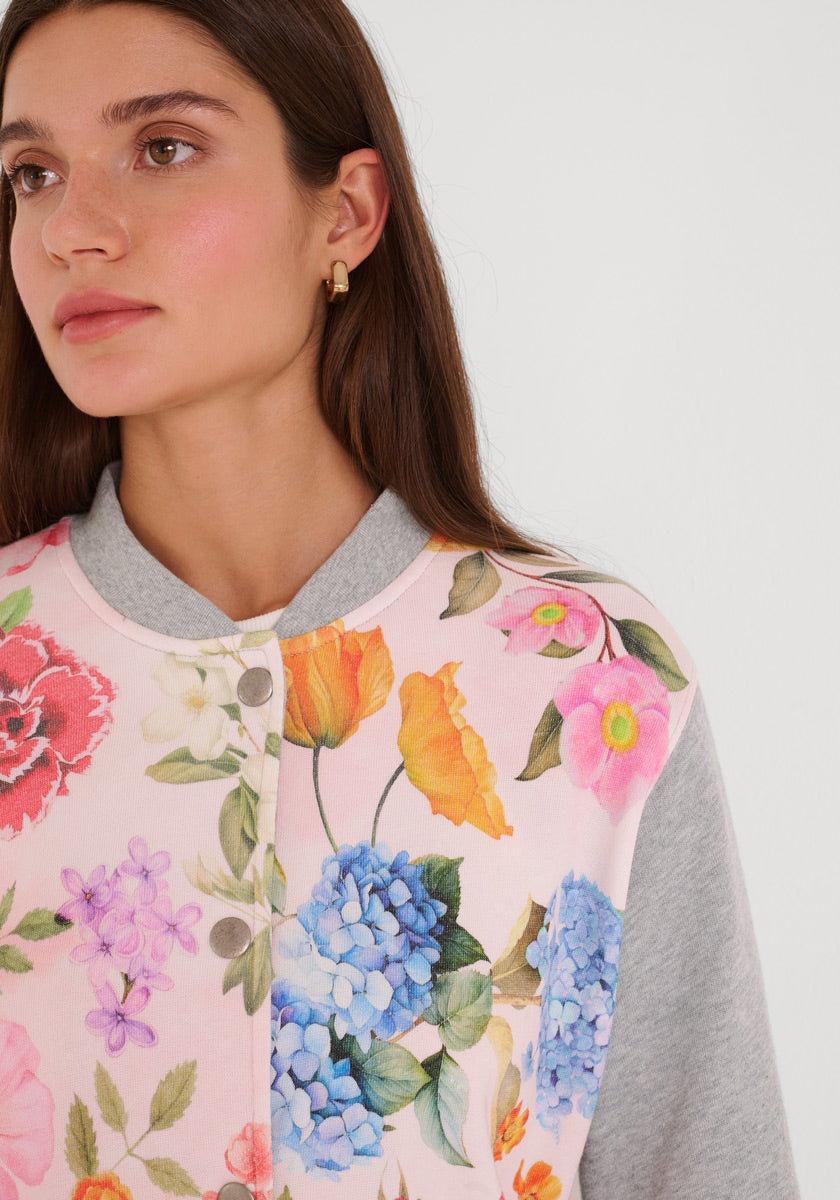 Eine Frau mit glattem braunem Haar und goldenen Ohrringen trägt ein Princess Goes Hollywood-Knopfleistenoberteil im College-Stil mit einem blassrosa Grundton mit leuchtend blauen Hortensien, rosa und orangefarbenen Blüten, grünen Blättern und hellgrauem Kragen und Ärmeln. Sie blickt nach links.