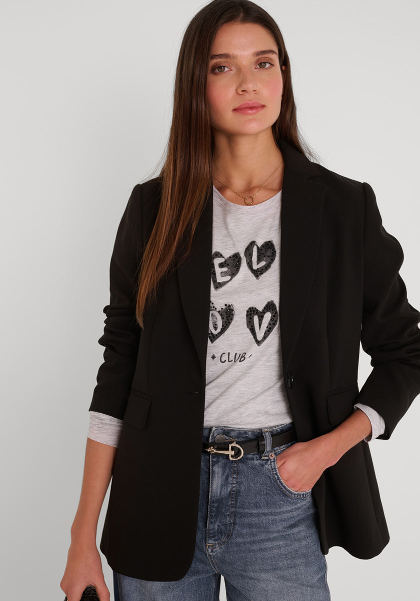 Eine Frau mit langen, glatten braunen Haaren trägt einen figurbetonten Blazer aus stretchigem Webstoff von Princess Goes Hollywood über einem hellgrauen Graphic-Tee mit schwarzen Herz-Designs und blauen Jeans. Sie blickt neutral nach vorne, die Hand in der Tasche.