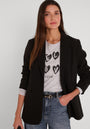 Eine Frau mit langen, glatten braunen Haaren trägt einen figurbetonten Blazer aus stretchigem Webstoff von Princess Goes Hollywood über einem hellgrauen Graphic-Tee mit schwarzen Herz-Designs und blauen Jeans. Sie blickt neutral nach vorne, die Hand in der Tasche.
