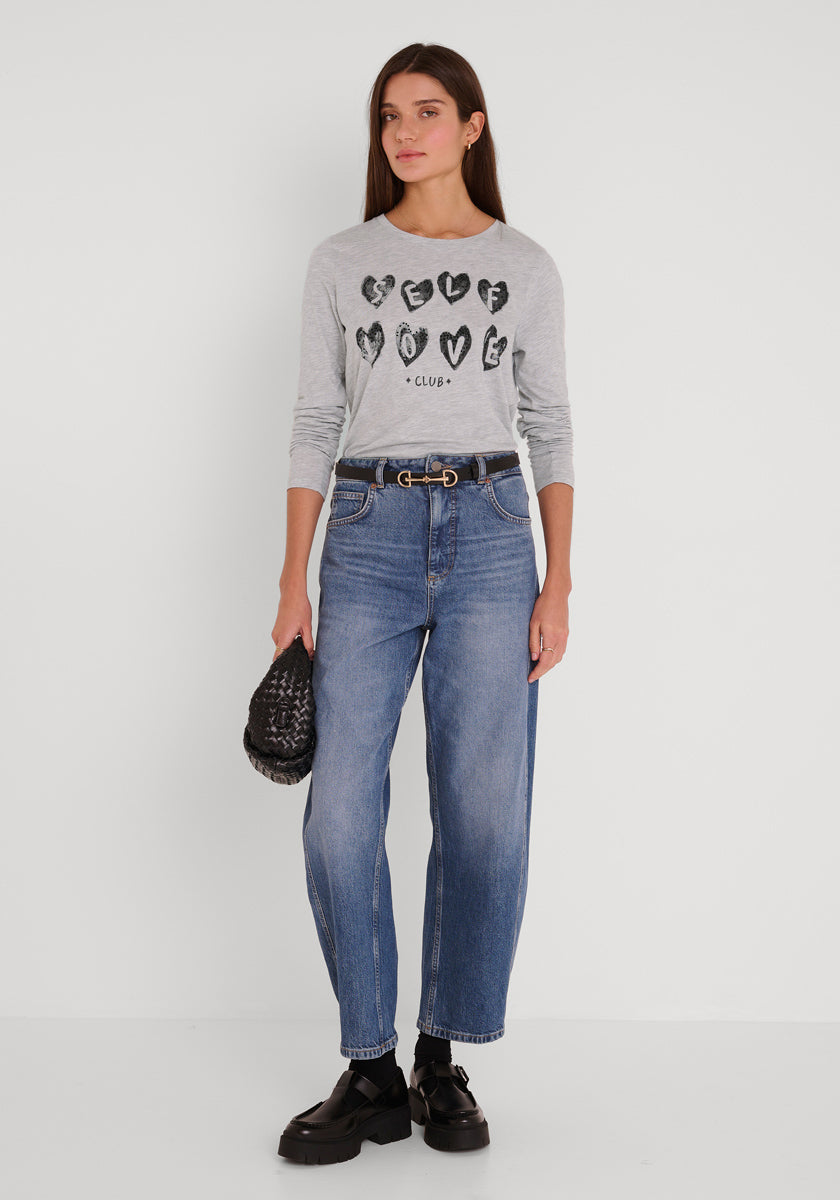 Eine Frau mit glattem braunem Haar und neutralem Gesichtsausdruck trägt ein hellgraues langärmeliges Baumwollhemd von Princess Goes Hollywood mit schwarzer Herzgrafik und dem Text "Self Love Club", blaue Jeans mit hoher Taille, klobige schwarze Schuhe und eine strukturierte Clutch.