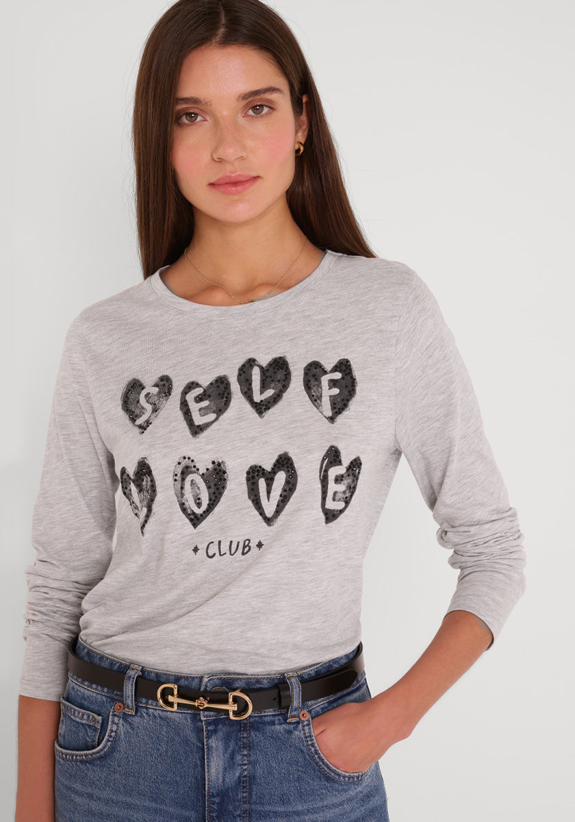 Eine Frau mit langen braunen Haaren trägt ein hellgraues langärmeliges Baumwollhemd von Princess Goes Hollywood mit der Aufschrift SELF LOVE CLUB" in schwarzen, herzförmigen Buchstaben, dazu blaue Jeans und einen schwarzen Gürtel vor einem schlichten weißen Hintergrund.