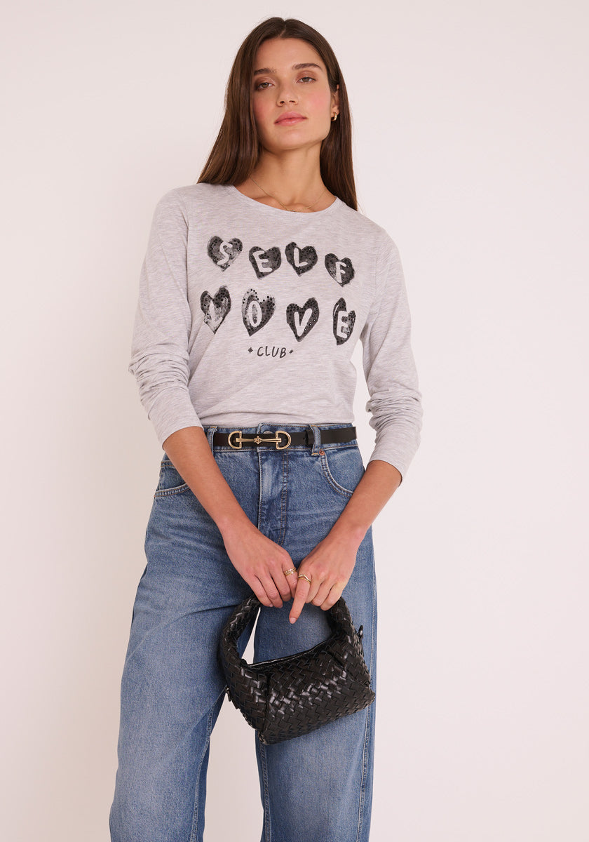 Eine Frau mit langen braunen Haaren trägt ein hellgraues Princess Goes Hollywood Baumwolle Langarmshirt mit der Aufschrift "SELF LOVE CLUB" in schwarzer Schrift, das in eine hochgekrempelte Bluejeans gesteckt ist und mit einem schwarzen Gürtel und einer gewebten Handtasche verziert ist.
