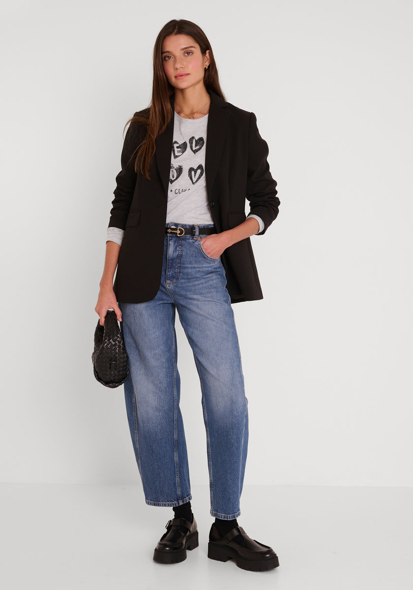 Eine Frau mit glattem braunem Haar steht vor einer weißen Wand. Sie trägt einen schwarzen Blazer über einem langärmeligen Baumwollhemd mit schwarzen Herzen von Princess Goes Hollywood Self Love Club, hochtaillierte Bluejeans, klobige Schuhe und eine gewebte Handtasche.