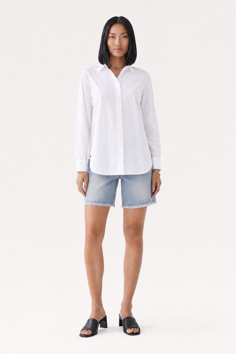 Eine Frau mit glattem schwarzem Haar trägt eine langärmelige weiße Hemdbluse aus Baumwolle von Princess Goes Hollywood, hellblaue Jeansshorts und schwarze Sandalen mit Absatz. Sie steht mit dem Blick nach vorne vor einem schlichten weißen Hintergrund, die Arme entspannt an den Seiten, und dezente Herzdetails verleihen ihrem Look eine charmante Note.
