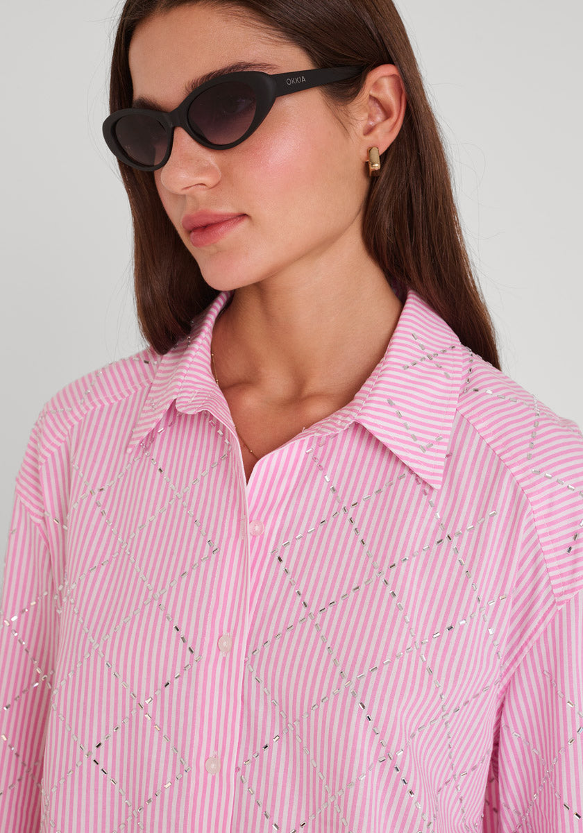 Eine Frau mit langen braunen Haaren trägt eine schwarze ovale Sonnenbrille, kleine goldene Ohrringe und ein rosa-weiß gestreiftes Baumwollhemd mit einem geometrischen Paillettenmuster von Princess Goes Hollywood. Sie blickt nach links vor einem schlichten hellen Hintergrund.